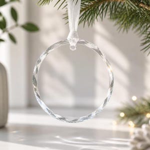 May include: A clear, circular glass ornament with a faceted, light-reflecting surface. The ornament is suspended by a white satin ribbon attached to a small metal loop at the top, creating a simple, transparent ring design for hanging.