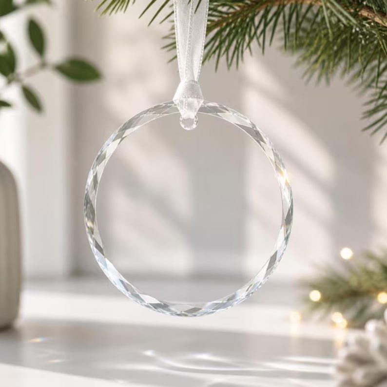 May include: A clear, circular glass ornament with a faceted, light-reflecting surface. The ornament is suspended by a white satin ribbon attached to a small metal loop at the top, creating a simple, transparent ring design for hanging.