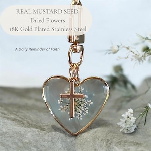 May include: A heart-shaped pendant featuring real mustard seeds and dried flowers encased in clear resin with an 18K gold-plated stainless steel frame and a small cross charm. The text reads: REAL MUSTARD SEED, Dried Flowers, 18K Gold Plated Stainless Steel, A Daily Reminder of Faith.