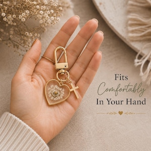 May include: A gold-toned keychain featuring a heart-shaped pendant filled with pressed white flowers and a small cross charm. The text on the right reads: Fits Comfortably In Your Hand. The keychain has a lobster clasp and a circular ring attachment.