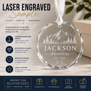 May include: A circular K9 crystal glass ornament with beveled edges, suspended by a white ribbon. The glass features a laser-engraved mountain and pine tree landscape above the text JACKSON Adventures EST. 2024 and two crossed arrows.