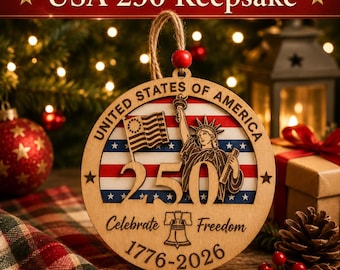 May include: A round wooden ornament featuring the Statue of Liberty, an American flag, and the number 250. The text reads United States of America, Celebrate Freedom, and 1776-2026. It includes a jute hanging loop with a red wooden bead.