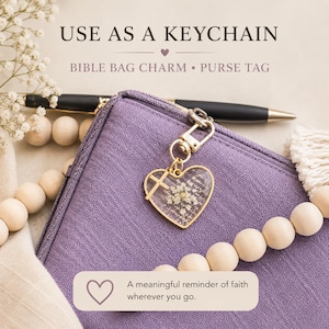 May include: A heart-shaped charm with a gold-tone metal frame containing pressed white flowers and a small cross, attached to a purple fabric bag. Text reads: USE AS A KEYCHAIN, BIBLE BAG CHARM, PURSE TAG, A meaningful reminder of faith wherever you go.