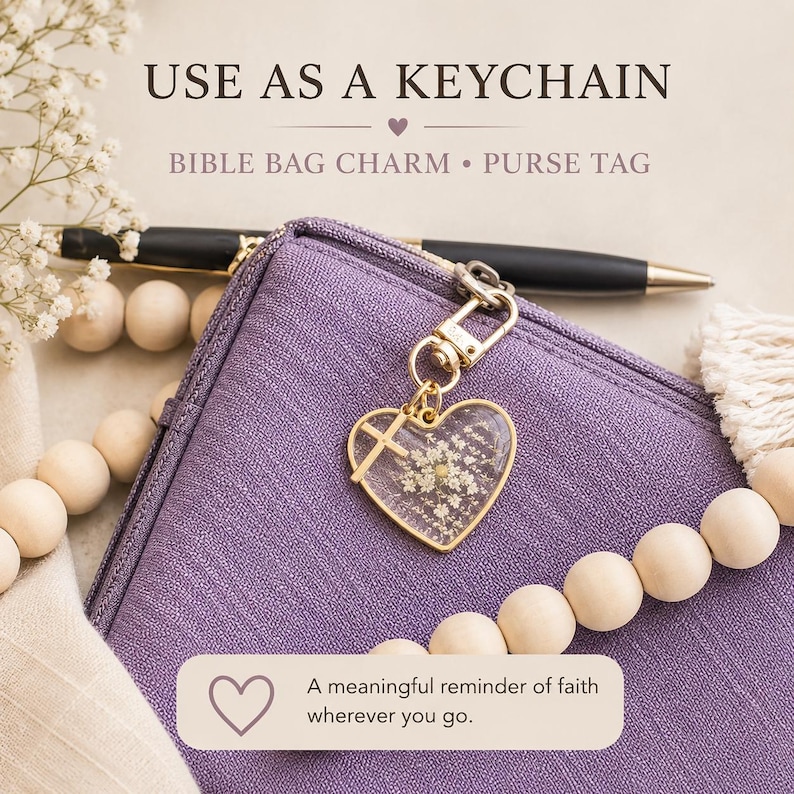 May include: A heart-shaped charm with a gold-tone metal frame containing pressed white flowers and a small cross, attached to a purple fabric bag. Text reads: USE AS A KEYCHAIN, BIBLE BAG CHARM, PURSE TAG, A meaningful reminder of faith wherever you go.
