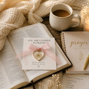 Może przedstawiać: Otwarta Biblia, dziennik modlitw i kubek kawy ułożone na drewnianej powierzchni. Obecna jest r&oacute;wnież karta z napisem "YOU ARE COVERED IN PRAYER" oraz zawieszka w kształcie serca z suszonymi kwiatami. Miękki, kremowy koc znajduje się w tle.