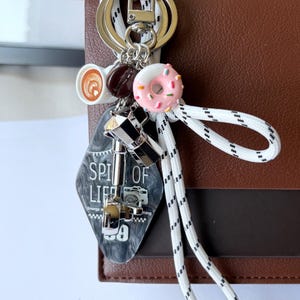 May include: A brown leather bag with a silver keyring holding charms. Charms include a coffee cup, a donut, and a black tag with the words "SPI OF LIFE" and a camera illustration. A white and black rope is attached.