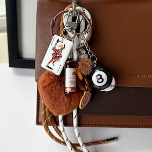 May include: A brown leather handbag with a decorative keychain. The keychain includes a fluffy brown heart, a miniature bottle, a playing card, and a black and white 8-ball charm. The bag has a gold-colored clasp.
