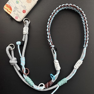 Könnte beinhalten: Eine Handyhülle mit einem Sonnensystem-Design und einem passenden Schlüsselband. Das Schlüsselband ist grau, blau und braun, mit Metallringen und dekorativen Knoten. Die Handyhülle ist cremefarben mit einer farbenfrohen Sonnensystem-Grafik.