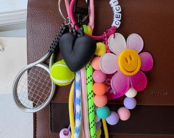 May include: A selection of vibrant keychains and charms, featuring a black heart, a tennis racket with a tennis ball, a flower with a smiley face, and beads. The word "CECI" is spelled out on white letter beads.