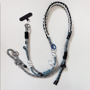 Puede incluir: Un cordón para teléfono con estampado en blanco, negro y gris con un agarre para teléfono negro. El cordón está hecho de cuerda trenzada y tiene un bucle para sujetarlo a una funda de teléfono. El cordón mide aproximadamente 76 cm de largo.