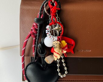 May include: A brown leather handbag with a gold clasp and a collection of charms. The charms include a black heart, a golf ball, a small dog figurine, and a red lobster. The word "HAPPY" is spelled out with white letter beads.