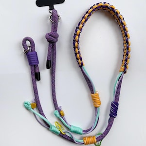 Op de afbeelding: Een veelkleurige telefoonlanyard met een gevlochten paarse en gouden nekband. De lanyard bevat paarse, turquoise en gouden accenten, met een metalen ring en een zwart bevestigingsstuk. Het ontwerp bevat knopen en decoratieve elementen.