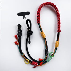 Può includere: Un cordino per telefono multicolore con un cinturino intrecciato rosso. Il cordino presenta cordoncini neri, verdi e arancioni, accentuati da dettagli gialli e rossi. Include un accessorio per telefono nero.