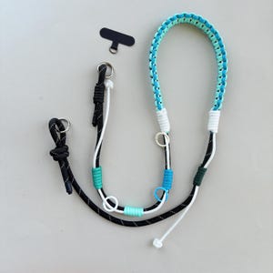 May include: A multi-coloured phone lanyard in black, white, and shades of blue and green. The lanyard has a black phone connector and a silver ring. The design includes a loop for wearing around the neck or wrist.