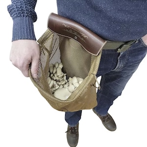 Könnte beinhalten: Ein brauner Ledergürtel mit einer braunen Netztasche. Die Tasche ist mit weißen Pilzen gefüllt und hat den Text "Ich bin ein Pilzjäger" auf dem Lederriemen.