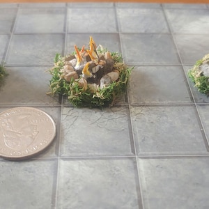 May include: Three miniature campfires with orange flames, surrounded by brown rocks and green moss. The campfires are different sizes, with the largest one in the back and the smallest one in the front. The campfires are sitting on a gray and white checkered surface.