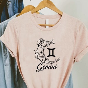Puede incluir: Una camiseta rosa claro con un gr&aacute;fico negro del signo zodiacal G&eacute;minis dentro de una luna creciente con detalles florales. El texto "Gemini" est&aacute; escrito debajo del gr&aacute;fico.