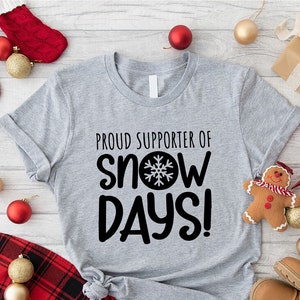 May include: A gray t-shirt with the text "Proud Supporter of Snow Days!" in black with a snowflake graphic. The shirt is surrounded by Christmas decorations, including red and gold ornaments, a gingerbread man, and a red star.