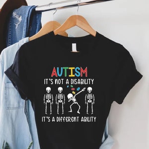 May include: Black t-shirt with a white skeleton dabbing and the text "AUTISM IT'S NOT A DISABILITY IT'S A DIFFERENT ABILITY". The skeleton is holding a puzzle piece with the colors of the autism awareness ribbon.