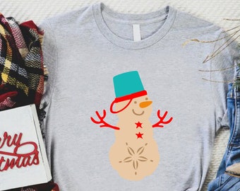 Könnte beinhalten: Ein graues T-Shirt mit einem Schneemann-Motiv. Der Schneemann trägt einen blauen Hut und einen roten Schal. Der Schneemann hat drei rote Sterne auf seinem Körper. Das T-Shirt liegt auf einer weißen Oberfläche mit einem karierten Schal, einer Jeans und einem Paar braunen Stiefeln.