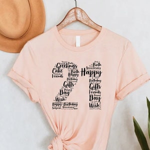 May include: A pink t-shirt with a black number 21 design made of words related to birthdays, such as "Happy", "Birthday", "Cake", "Gifts", and "Friends".