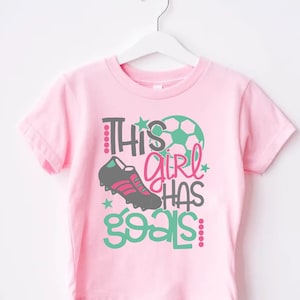May include: Pink t-shirt with a green, black, and white soccer ball graphic and the text "This girl has goals".