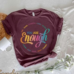May include: A maroon t-shirt with a colorful graphic that says "You Are Enough" surrounded by positive affirmations. The affirmations are "Kind, Tough, Powerful, Loved, Valued, Strong, Bold, Brave, Smart, Capable".