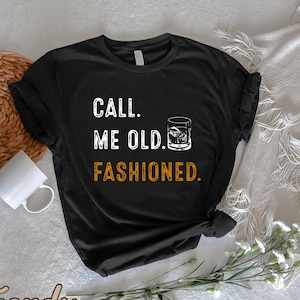 May include: Black t-shirt with the text "CALL. ME OLD. FASHIONED." in white and orange lettering. A graphic of a cocktail glass is included. The shirt is displayed on a neutral surface with a white mug and decorative elements.
