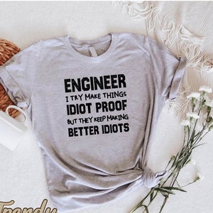 May include: A light gray t-shirt with the text "ENGINEER I TRY MAKE THINGS IDIOT PROOF BUT THEY KEEP MAKING BETTER IDIOTS" in black. The shirt is knotted at the bottom. The brand name "Trendy CUSTOMGOODS" is at the bottom left.