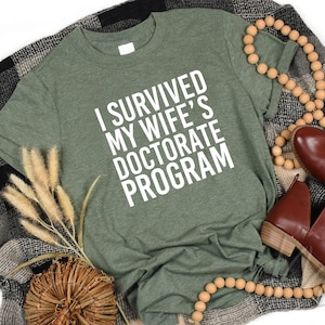 May include: A green t-shirt with the text "I survived my wife's doctorate program" in white letters.