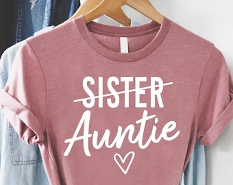 May include: A dusty rose t-shirt with the word "Sister" crossed out and replaced with "Auntie" in white script. A small heart is below the word "Auntie". The shirt is knotted at the bottom and hangs on a wooden hanger.