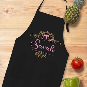 Puede incluir: Delantal negro con un dise&ntilde;o rosa y dorado que presenta un secador de pelo, tijeras y peine, con el nombre "Sarah" en escritura rosa. El delantal tiene dos bolsillos y se exhibe sobre una superficie de madera con una pi&ntilde;a, tomate y pimiento verde.