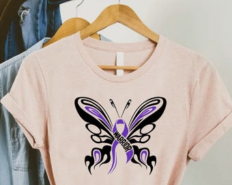 Może przedstawiać: Jasnoróżowy t-shirt z czarnym wzorem motyla. Motyl ma fioletową wstążkę z napisem "Warrior".