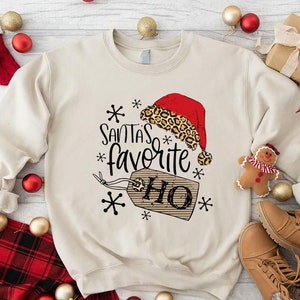 May include: A beige crewneck sweatshirt featuring a graphic print of a red Santa hat with a leopard print trim, the text "SANTA'S favorite HO" in black script and block lettering, and a brown gift tag, surrounded by black snowflake icons.