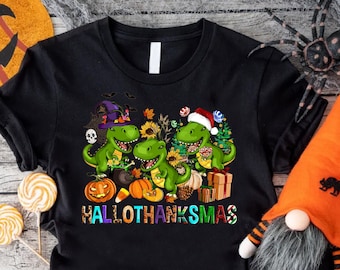 Könnte beinhalten: Schwarzes T-Shirt mit einem Motiv von drei Dinosauriern, die sich für Halloween, Thanksgiving und Weihnachten verkleidet haben. Der Text "Hallothanksmas" ist unter den Dinosauriern gedruckt.