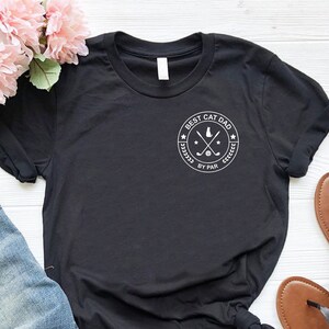 May include: Black t-shirt with the text "BEST CAT DAD BY PAR" in a white circular design. The shirt is paired with blue jeans and brown sandals. Pink flowers are in the background.