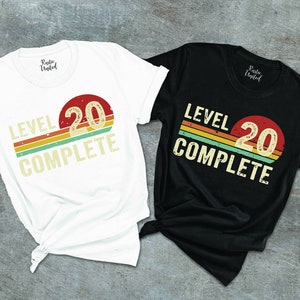 May include: Two t-shirts with a retro design. The white t-shirt has the text "Level 20 Complete" with a red, yellow, orange, and green striped design. The black t-shirt has the same design but in black.