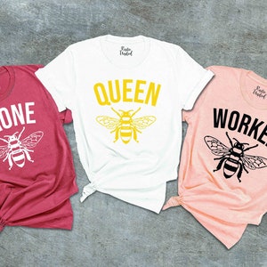 May include: Three t-shirts with bee designs and text. The red shirt says "Drone" with a white bee. The white shirt says "Queen" with a yellow bee. The pink shirt says "Worker" with a black bee.