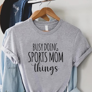 May include: Gray t-shirt with the text "Busy Doing Sports Mom Things" in black lettering.