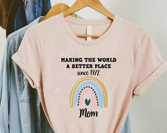 Peut inclure: Un t-shirt rose avec un motif arc-en-ciel et le texte "Making the world a better place since 1972 Mom".