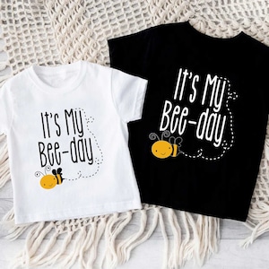 May include: Two white short-sleeved t-shirts with black text and a cartoon bee graphic. The text on the shirts reads "It's My Bee-day".