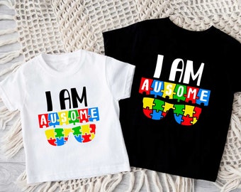 May include: Two short-sleeved t-shirts, one white and one black, with the text "I AM AUSOME". The word "AUSOME" is designed as sunglasses made of colorful puzzle pieces. The shirts are likely for children.