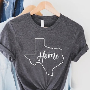May include: A dark charcoal gray short-sleeve t-shirt featuring a white outline of the state of Texas with the word Home written in a white cursive script font across the center of the state shape. The shirt has a crew neckline and rolled sleeves.