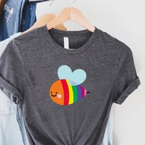 May include: Dark gray t-shirt featuring a colorful cartoon bee with rainbow stripes and blue heart-shaped wings. The shirt is displayed on a wooden hanger, with a light blue denim shirt and a brown hat in the background.