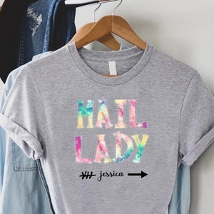 May include: Grey t-shirt with a tie-dye design featuring the text "Mail Lady" and the hashtag "#jessica" with an arrow.
