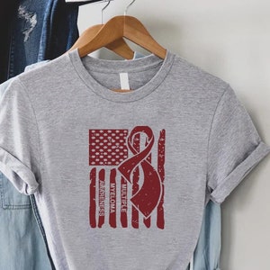 May include: Gray t-shirt with a red distressed American flag design. The flag has a red ribbon with the words "Multiple Myeloma Awareness" printed on it.