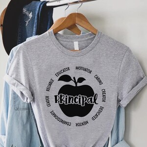May include: A heather gray short-sleeve t-shirt featuring a black graphic of an apple with the word Principal written across the center. Surrounding the apple are the words Reliable, Educator, Motivator, Caring, Creator, Dedicated, Mentor, Compassionate, and Leader arranged in a circular pattern.