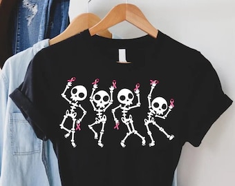 Puede incluir: Camiseta negra con un gráfico de cuatro esqueletos sosteniendo cintas rosadas. Los esqueletos están bailando y sonriendo.
