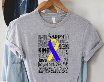 May include: A grey t-shirt featuring a Down Syndrome Awareness design. The design includes a yellow and blue ribbon, with words such as "inspiring," "happy," and "love." The t-shirt is hanging on a wooden hanger.