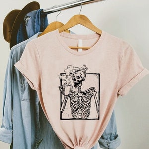 May include: A pink t-shirt with a black and white illustration of a skeleton holding a steaming mug. The skeleton is smiling and appears to be enjoying the drink.
